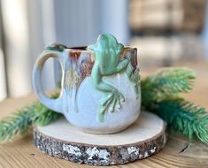 a green frog sitting on top of a white coffee cup next to a pine branch