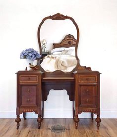 an antique wooden vanity with mirror and flowers on the top shelf, in front of a white wall