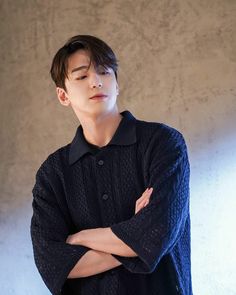 a young man with his arms crossed standing in front of a white wall wearing a black sweater