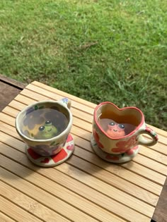 two coffee cups sitting on top of a wooden table next to each other in front of a grassy field