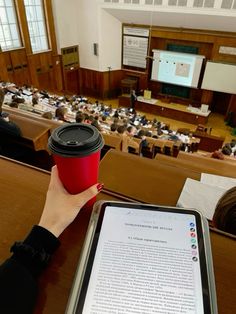 a person holding a red cup in front of an auditorium full of people sitting at desks