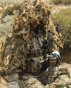 a man with long curly hair sitting on the ground in camouflage gear and holding a camera