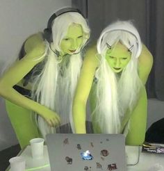 two women dressed in green and white are looking at a laptop screen with headphones on