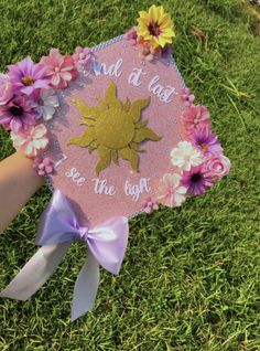 a hand holding a pink and gold graduation cap with flowers on the side, in grass