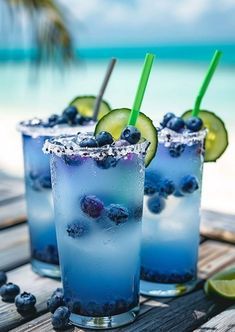 three glasses filled with blueberries and cucumber on a table near the ocean