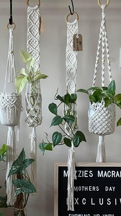 macrame plant holders hanging from hooks on a wall with a sign next to them