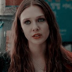 a woman with long brown hair standing in front of a building and looking at the camera