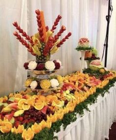 an arrangement of fruits and flowers on a table with white drapes in the background