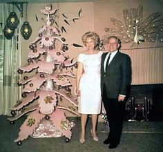 a man and woman standing next to a pink christmas tree in front of a wall