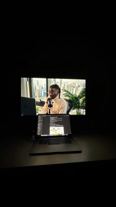 a laptop computer sitting on top of a desk in front of a tv screen with a man talking into a microphone