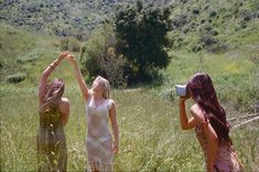 three women standing in tall grass with their hands up to the sky and one holding a camera