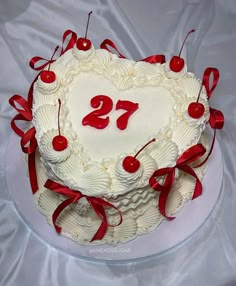 a heart shaped cake with red ribbons and cherries on it, sitting on a white table cloth