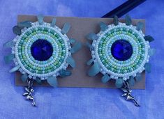 These green and blue hand beaded earrings are 2in in diameter. The center is a plastic blue gem and the beads are size 11 green, white, and blue seed beads. The edging is done with size 8 pearly white beads and aventurine chips. There is a stainless steel fairy charm hanging off the earring. Backed with deer hide and signed by me. Embroidery Flats, Fairy Earrings, Blue Beaded Earrings, Deer Hide, Fairy Charms, Blue Fairy, Handmade Earrings Beaded, Earrings Beaded, Blue Hand