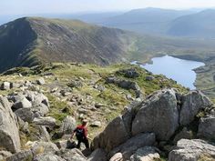 a man climbing up the side of a mountain on top of a rocky hill with a lake in the distance