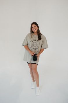 a woman standing in front of a white wall holding a camera and smiling at the camera