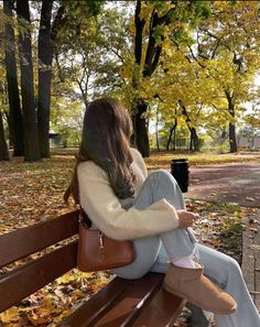 a woman sitting on top of a wooden bench next to trees filled with fall leaves