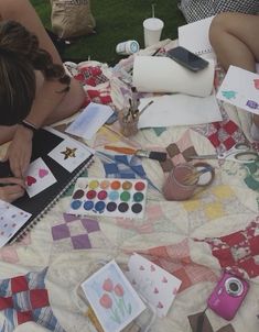 two women sitting on a blanket with art supplies and watercolors in front of them