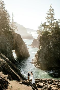 two people are standing on the rocks near the water and some trees in the background