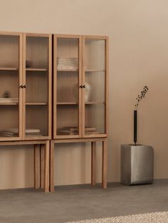 a tall wooden cabinet with glass doors next to a vase on the floor in front of a beige wall