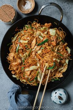 a wok filled with noodles and chicken on top of a table next to two wooden chopsticks