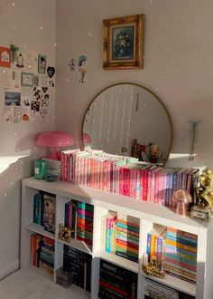 a white book shelf filled with lots of books next to a wall mounted round mirror