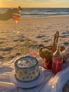 there is a cake on the beach next to some wine glasses and a basket with food