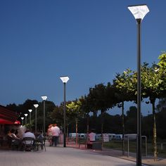 people are sitting at tables under street lamps on the sidewalk in front of some trees