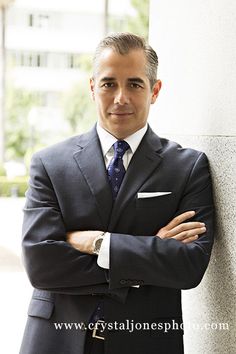 a man in a suit and tie leaning against a wall with his arms crossed, looking at the camera