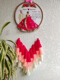 a wall hanging with a pink and white dress on it next to a potted plant