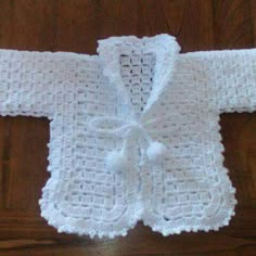 a white crocheted baby jacket with a bow on the front and back, sitting on a wooden floor