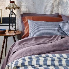 a bed with pillows and blankets on it next to a night stand in front of a window