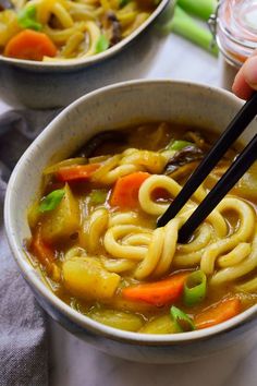 someone holding chopsticks over a bowl of noodles and vegetables with soup in the background