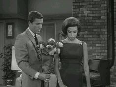 a man and woman standing next to each other in front of a brick wall holding flowers