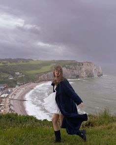 a woman standing on top of a lush green hillside next to the ocean and cliffs