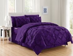 a bed with purple comforter and pillows in a room next to a white table