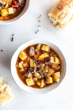 two bowls of stew next to rolls and bread on a table with utensils