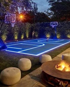 an outdoor basketball court is lit up at night with fire pit and seating around it