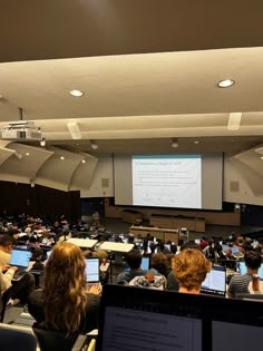 an auditorium full of people with laptops in front of them and a projector screen on the wall