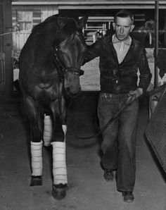 black and white photograph of man leading horse in stable