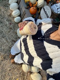 a woman is sitting on the ground with pumpkins and squash in the background, drinking from a cup