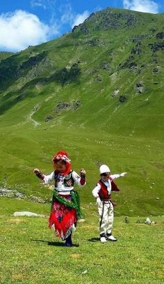 two people dressed in traditional clothing standing on a grassy field with mountains in the background