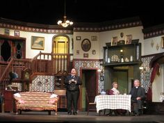 three people sitting at a table in front of a stage set with chairs and couches