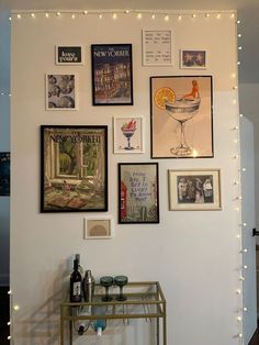 a wall covered in pictures and framed art with lights on the walls above it is a bar cart filled with drinks