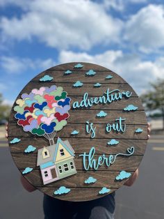 a person holding up a wooden sign that says adventure is out there with a house and balloons