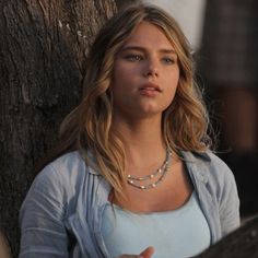 a woman standing next to a tree with her hand on her hip and looking at the camera