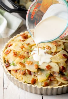 a person pouring milk into a pie with potatoes and bacon in it on a white wooden table