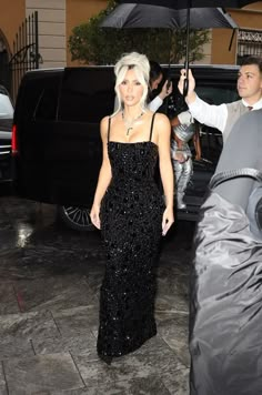 lady with white hair and black dress holding an umbrella in front of a man standing next to her