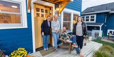 three people standing in front of a blue house with yellow flowers on the porch and door