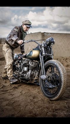 a man standing next to a motorcycle on top of a dirt field with clouds in the background