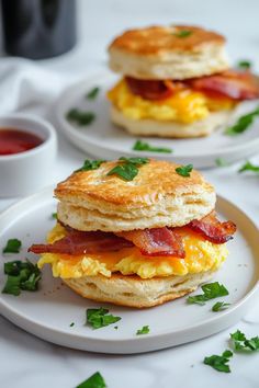 two breakfast sandwiches on plates with bacon and eggs